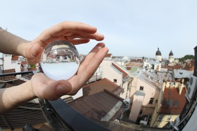 Photo of View of beautiful city street, overturned reflection. Man holding crystal ball outdoors, closeup. Wide-angle lens