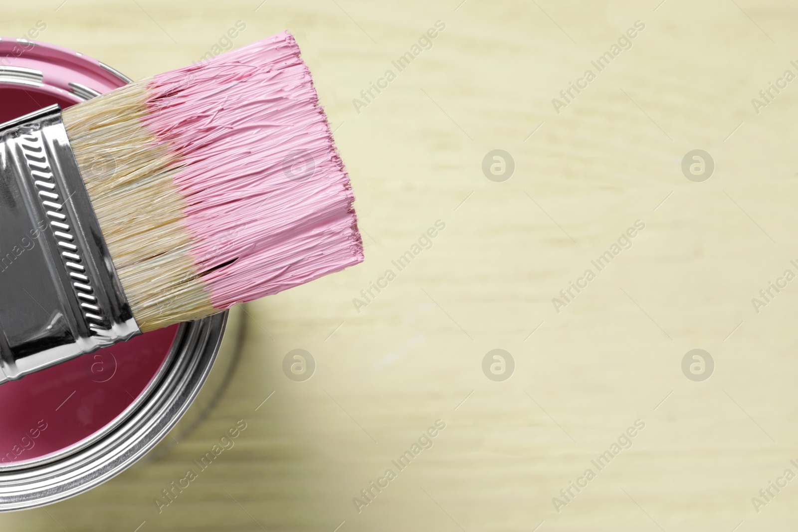 Photo of Can of pink paint and brush on pale yellow table, closeup. Space for text