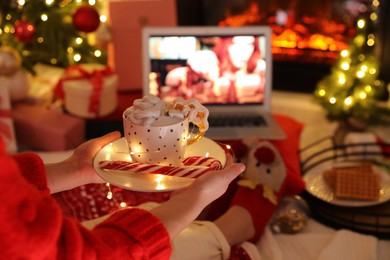 MYKOLAIV, UKRAINE - DECEMBER 23, 2020: Woman with sweet drink watching The Queen's Gambit series on laptop near fireplace at home, closeup. Cozy winter holidays atmosphere