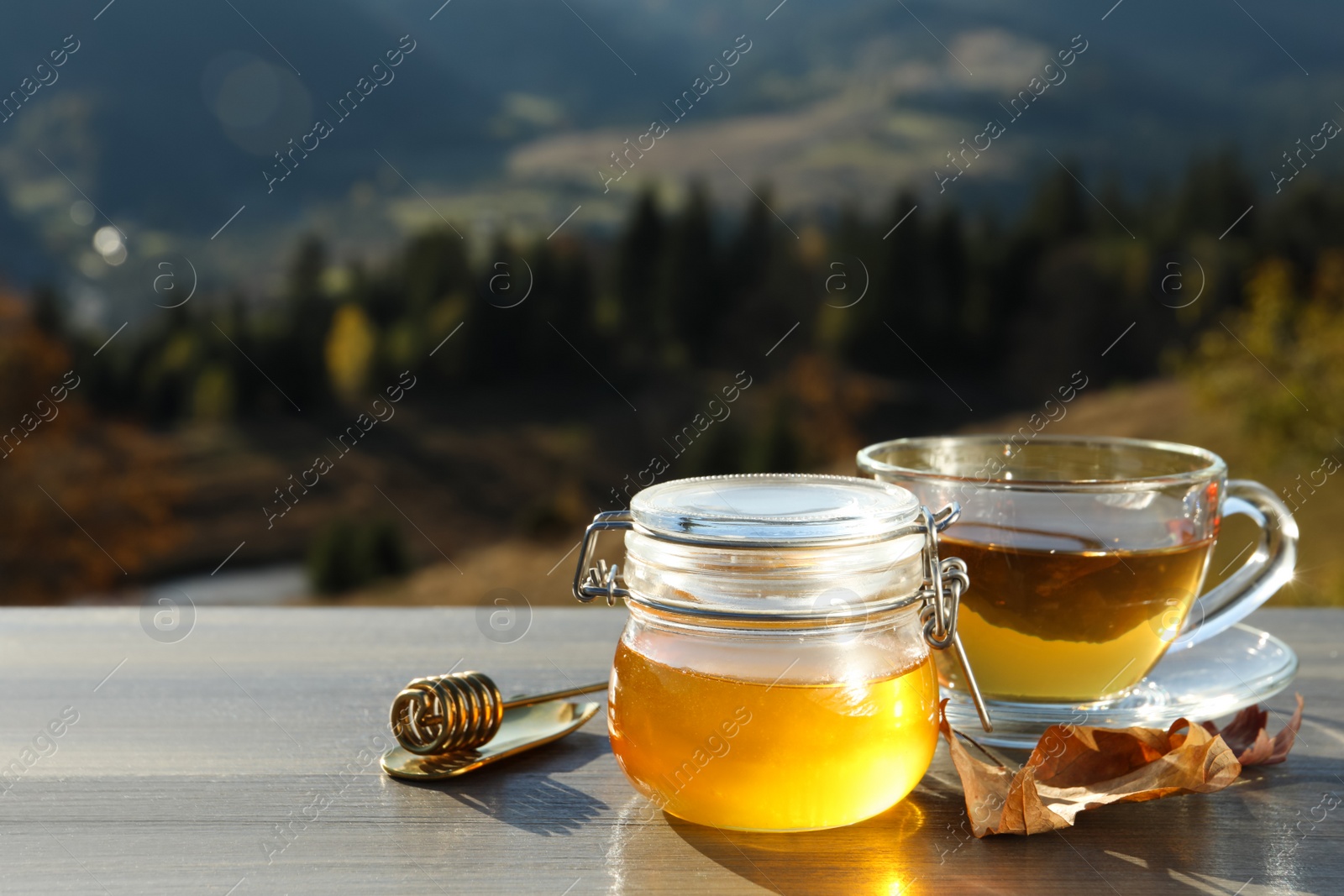 Photo of Fresh aromatic honey and cup of hot tea on grey wooden table against mountain landscape. Space for text