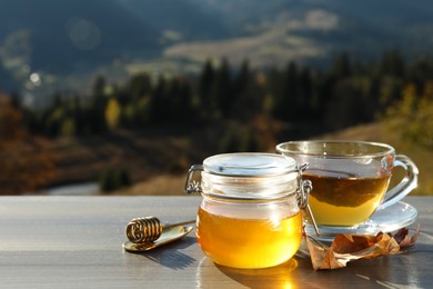 Photo of Fresh aromatic honey and cup of hot tea on grey wooden table against mountain landscape. Space for text