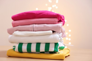 Photo of Stack of folded clothes on wooden table against beige background with blurred lights. Space for text