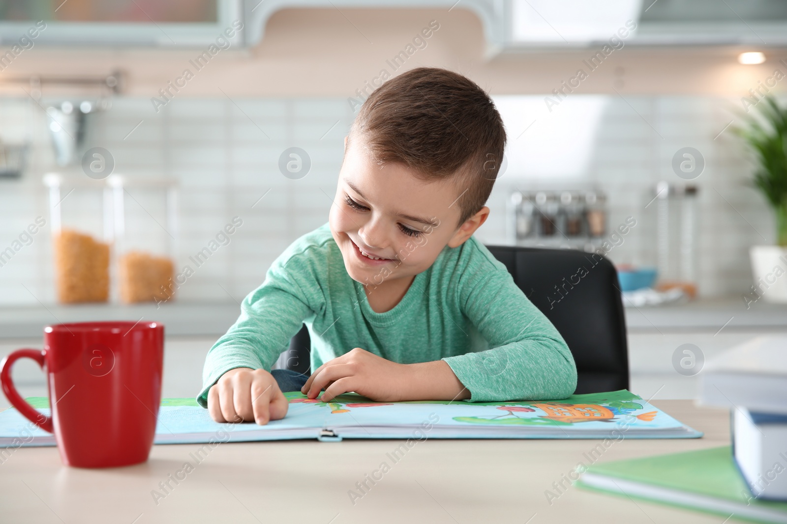 Photo of Cute little boy reading book at table in kitchen