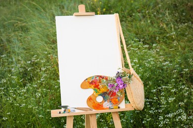 Photo of Wooden easel with blank canvas, painting equipment and flowers in meadow. Space for text