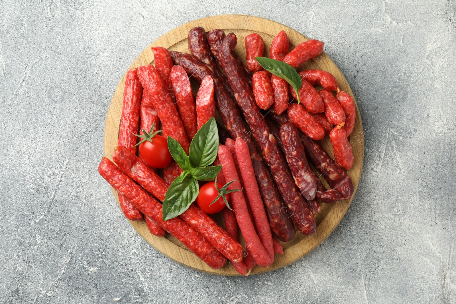 Photo of Different thin dry smoked sausages, basil and tomatoes on light grey table, top view