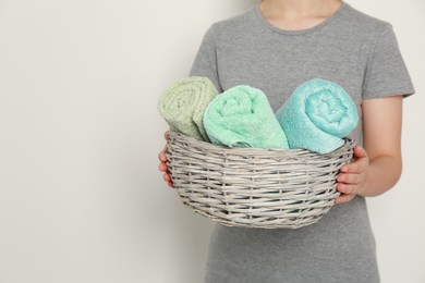 Photo of Woman holding wicker basket with rolled soft terry towels on light background, closeup. Space for text