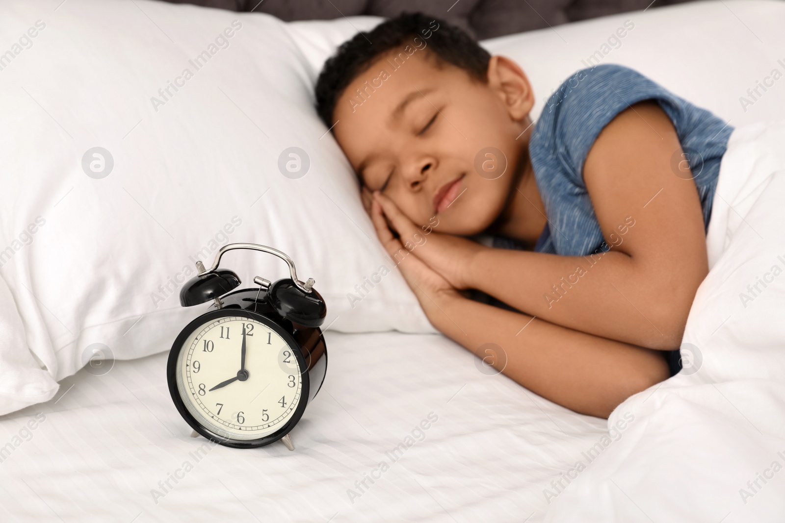 Photo of Cute little African-American boy with alarm clock sleeping in bed