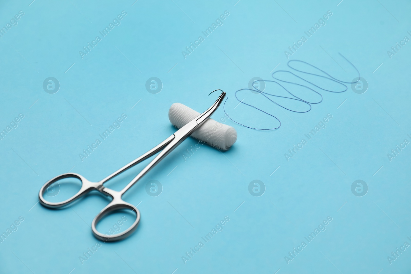 Photo of Forceps with suture thread and bandage roll on light blue background. Medical equipment