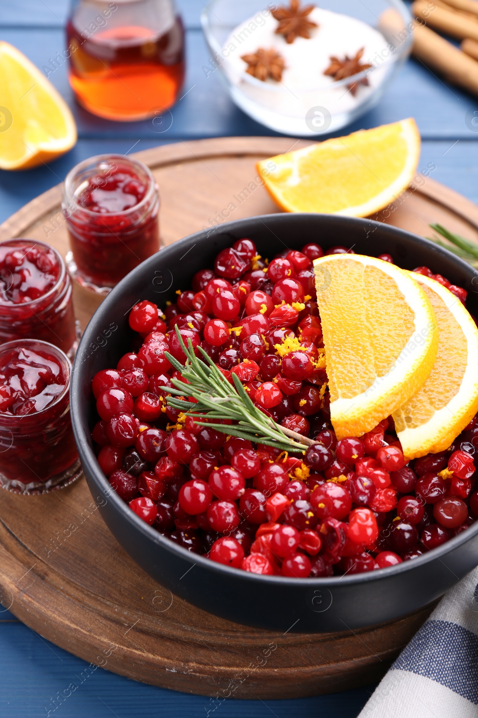 Photo of Cranberries in bowl, jars with sauce and ingredients on blue wooden table, closeup