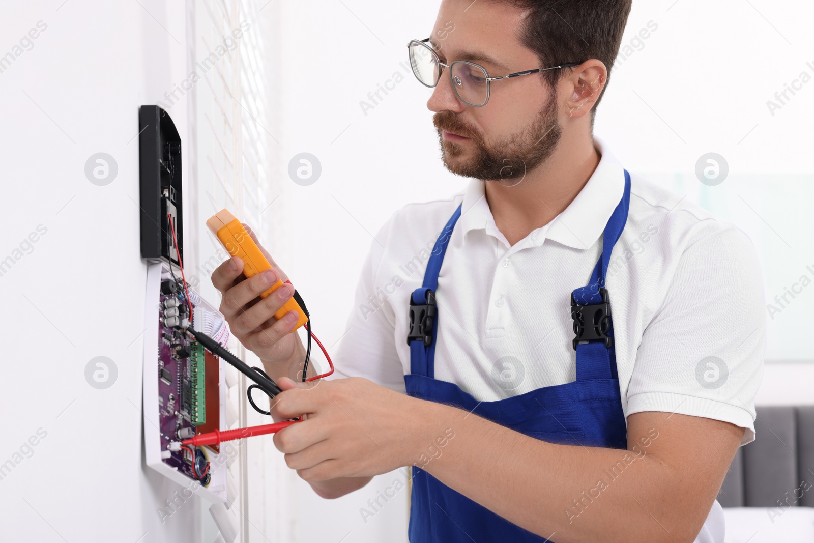 Photo of Technician using digital multimeter while installing home security alarm system on white wall indoors