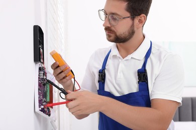 Technician using digital multimeter while installing home security alarm system on white wall indoors