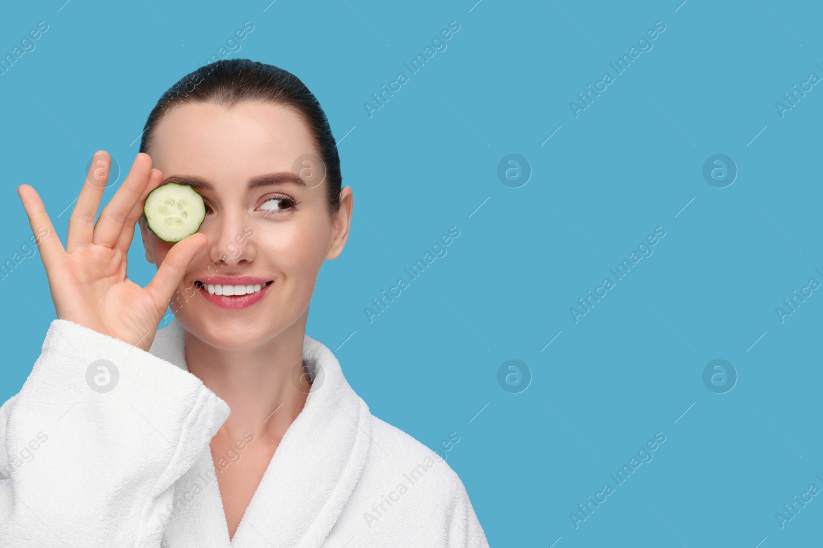 Photo of Beautiful woman covering eye with piece of cucumber on light blue background, space for text