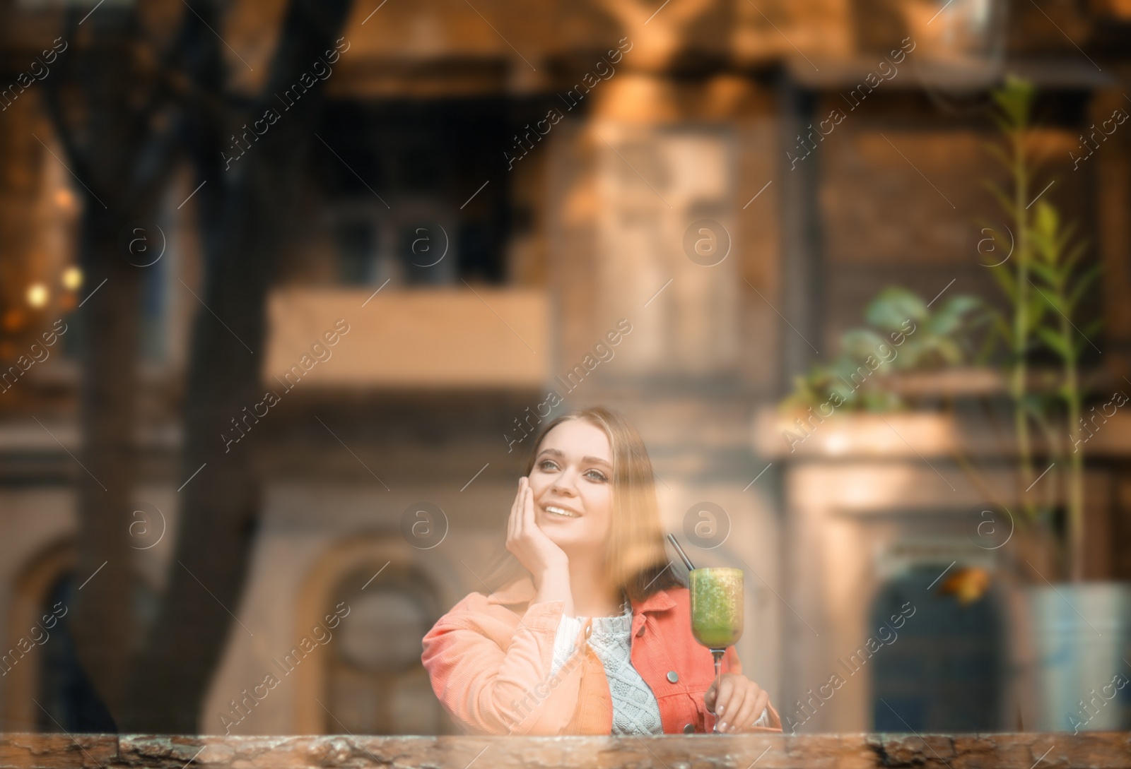 Photo of Pretty young woman with cocktail at table in cafe, view from outdoors through window