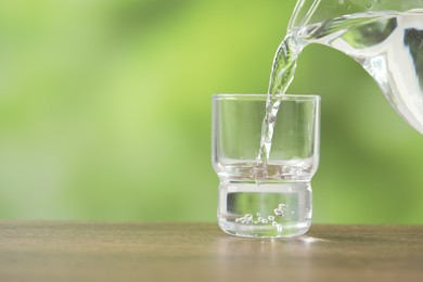 Pouring fresh water from jug into glass on wooden table against blurred green background, closeup. Space for text