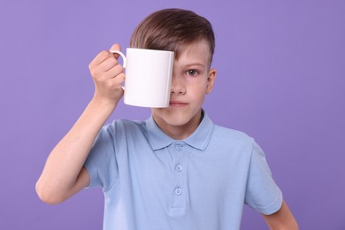 Cute boy covering eye with white ceramic mug on violet background