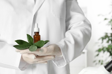 Photo of Lab assistant holding green twig and bottle of essential oil on blurred background, closeup. Plant chemistry