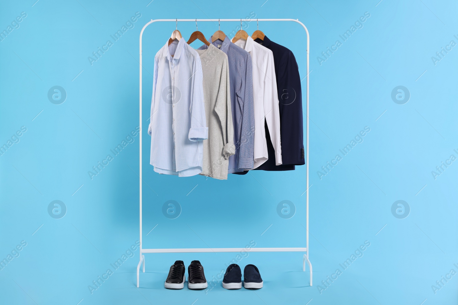 Photo of Rack with stylish clothes on wooden hangers and sneakers against light blue background