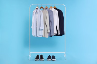Photo of Rack with stylish clothes on wooden hangers and sneakers against light blue background