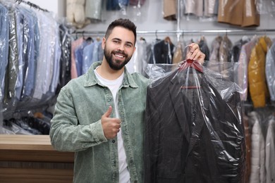 Photo of Dry-cleaning service. Happy man holding hanger with jacket in plastic bag and showing thumb up indoors