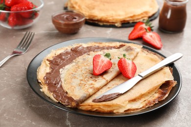 Photo of Delicious thin pancakes with chocolate paste and strawberries on grey marble table