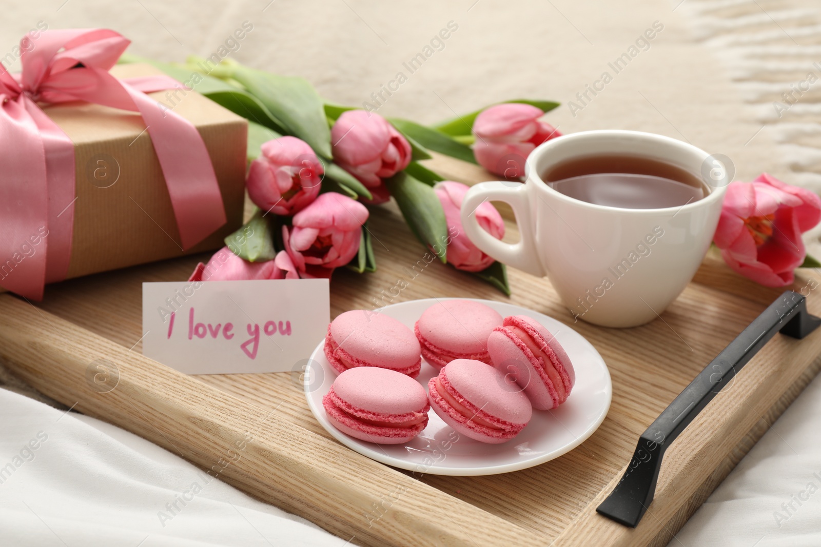 Photo of Tasty breakfast served in bed. Delicious macarons, tea, flowers, gift box and card with phrase I Love You on tray