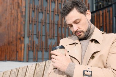 Tired man with cup of coffee sleeping while sitting on wooden bench outdoors. Space for text