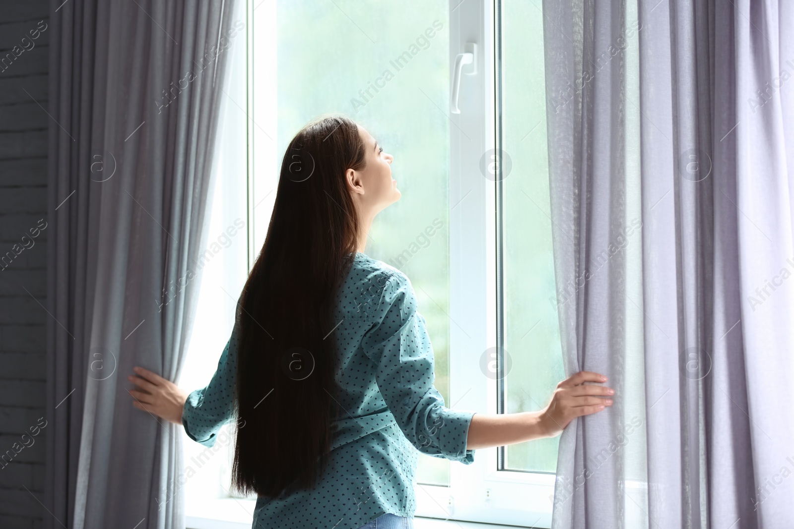 Photo of Young woman opening curtains in room near big beautiful window