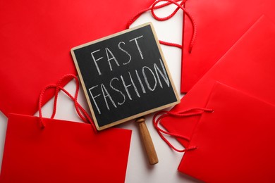 Photo of Small chalkboard with phrase Fast Fashion and red shopping bags on white background, top view