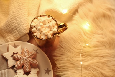 Photo of Cup of hot drink with marshmallows, cookies and Christmas lights on table, above view