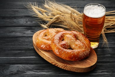 Photo of Tasty pretzels, glass of beer and wheat spikes on black wooden table