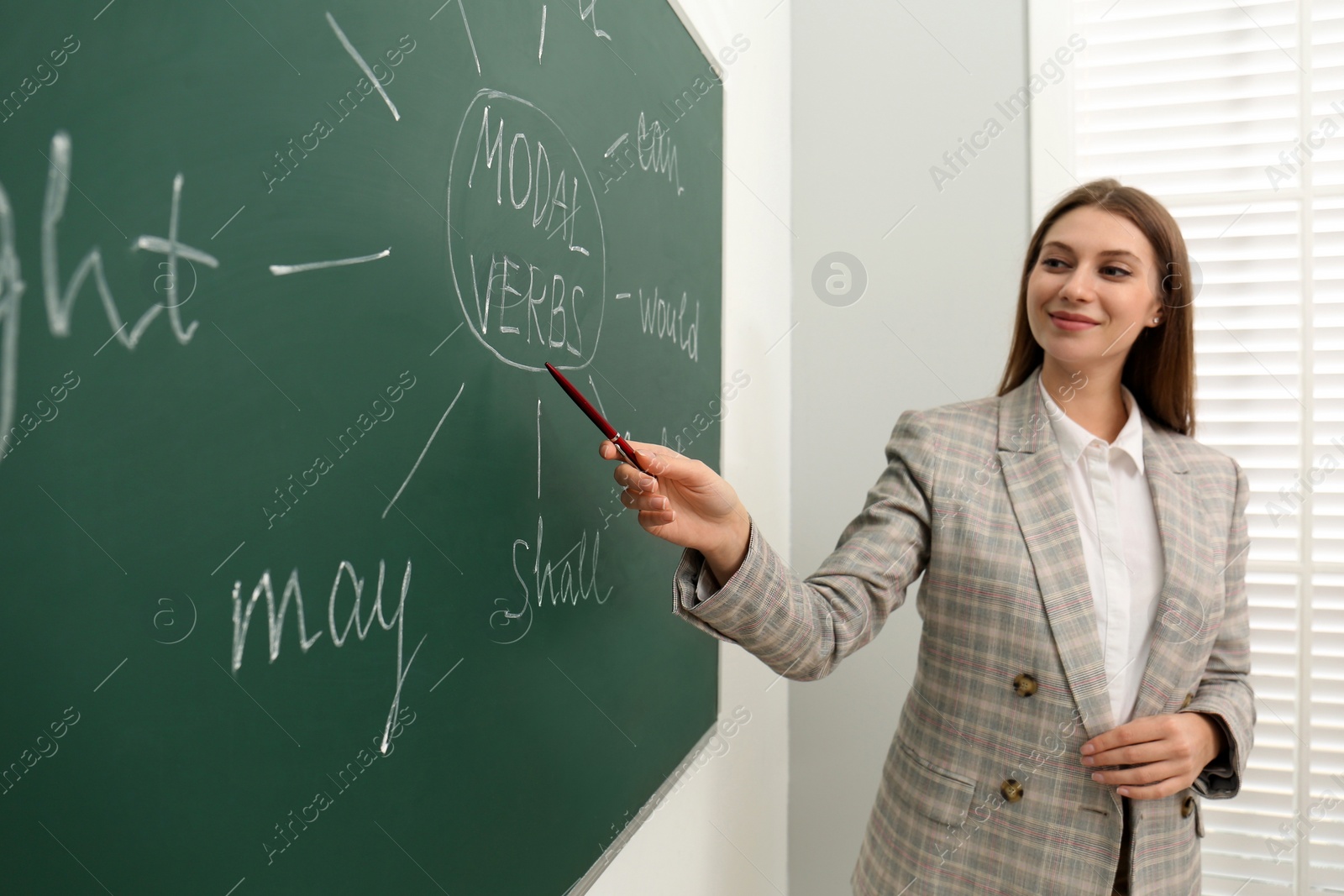 Photo of English teacher giving lesson on modal verbs near chalkboard in classroom