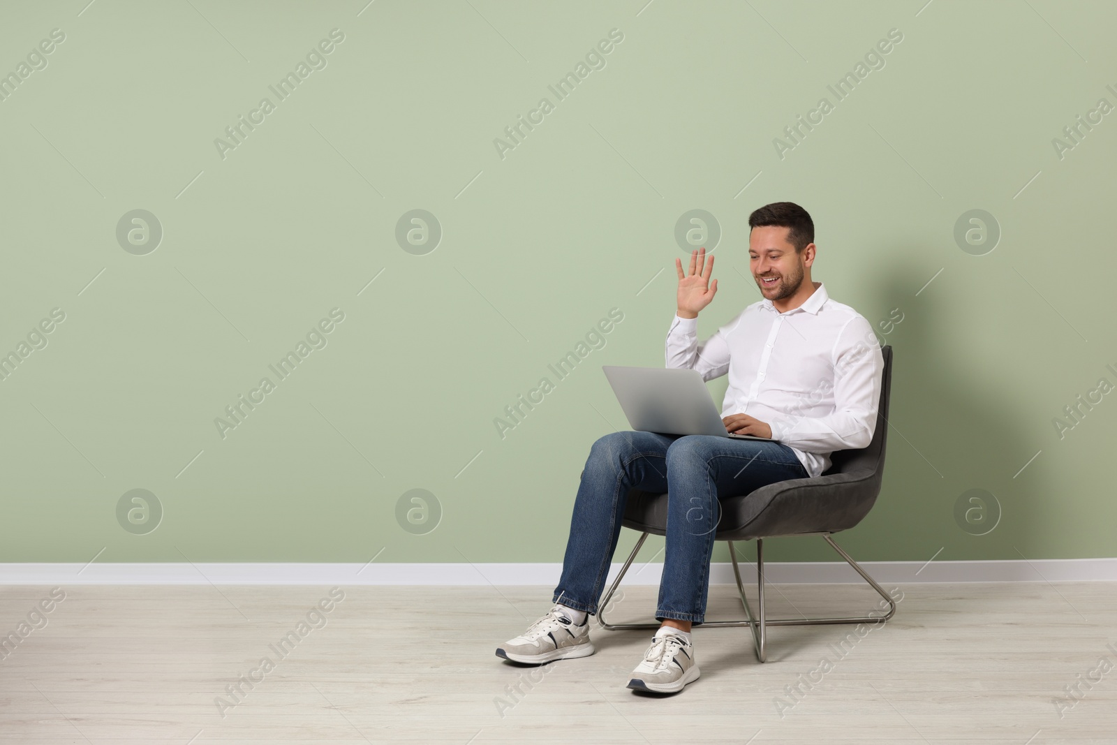 Photo of Happy man sitting in armchair and having video chat via laptop near light green wall indoors, space for text