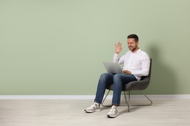 Happy man sitting in armchair and having video chat via laptop near light green wall indoors, space for text