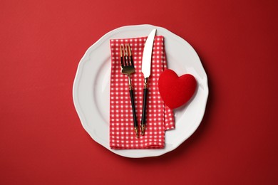 Plate with cutlery and decorative heart on red table for romantic dinner, top view