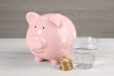 Photo of Water scarcity concept. Piggy bank, coins and glass of drink on white wooden table