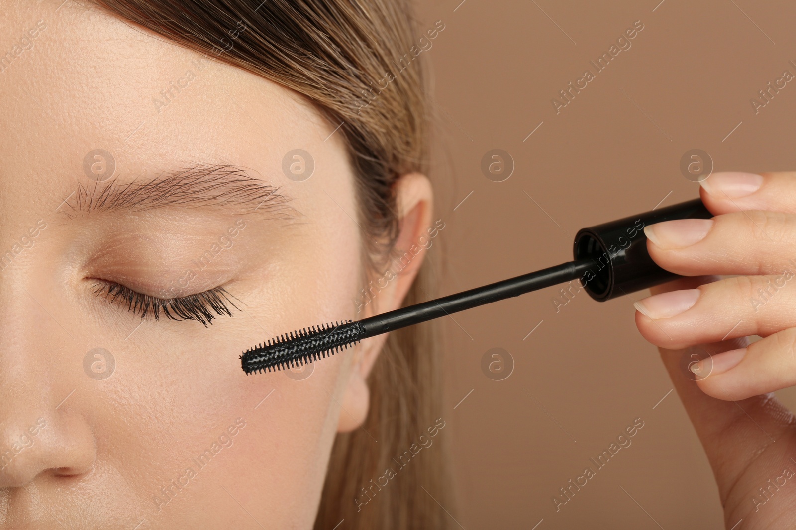 Photo of Woman applying mascara onto eyelashes against light brown background, closeup