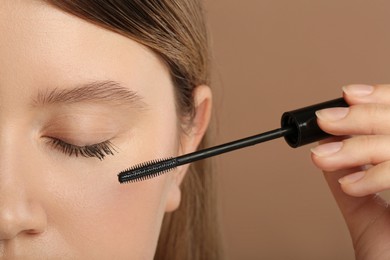 Woman applying mascara onto eyelashes against light brown background, closeup