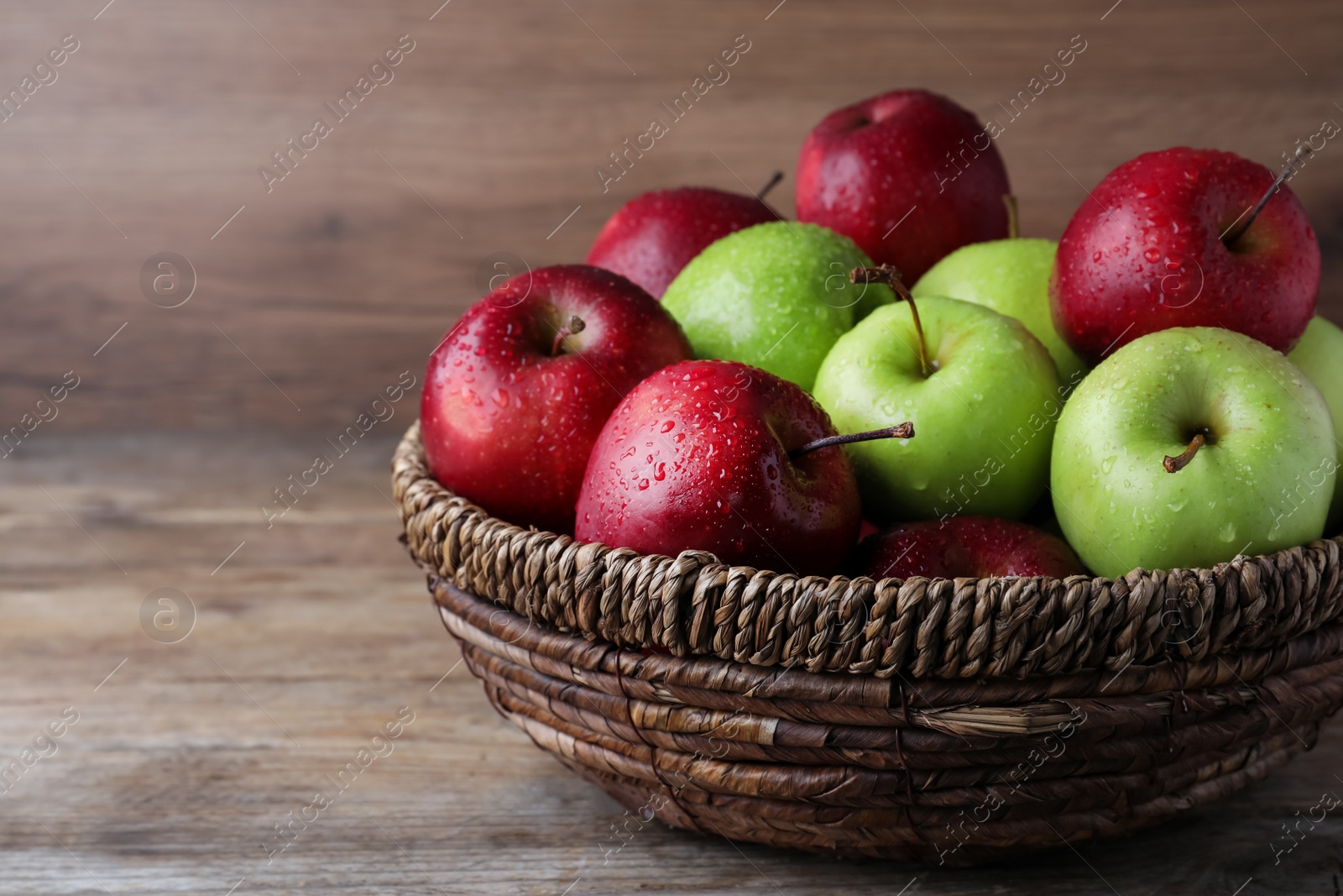 Photo of Fresh ripe green and red apples with water drops in wicker bowl on wooden table