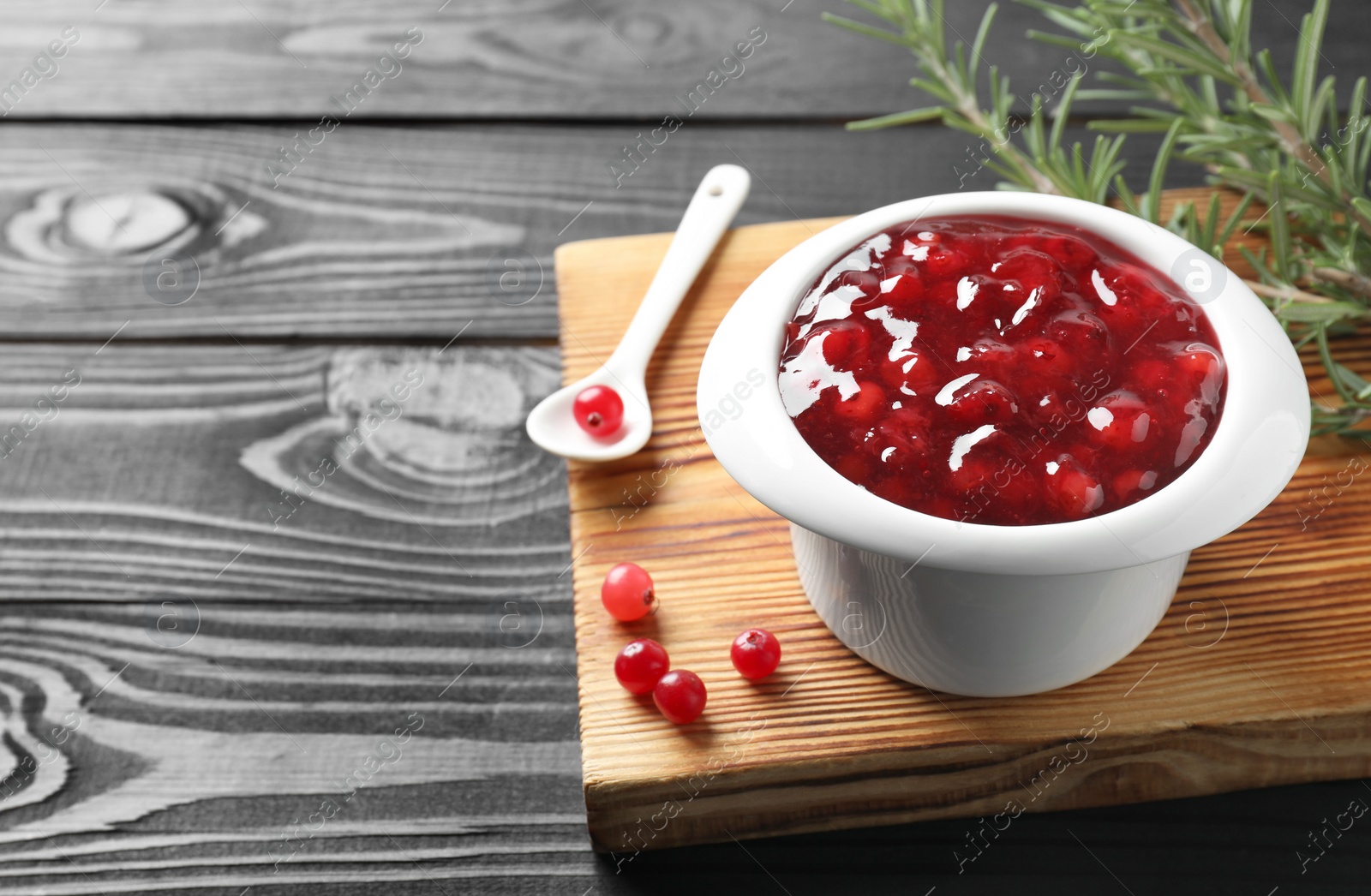 Photo of Cranberry sauce in bowl, fresh berries, spoon and rosemary on black wooden table, closeup. Space for text