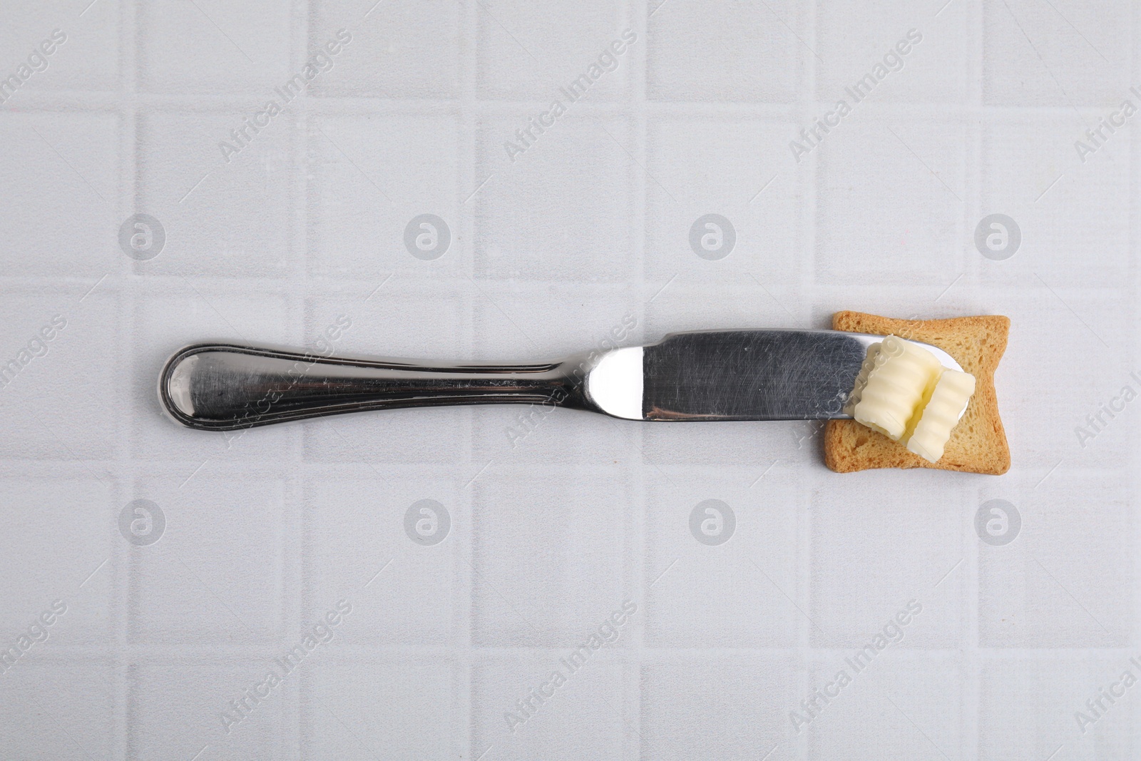 Photo of Tasty butter curl, knife and piece of dry bread on white tiled table, top view