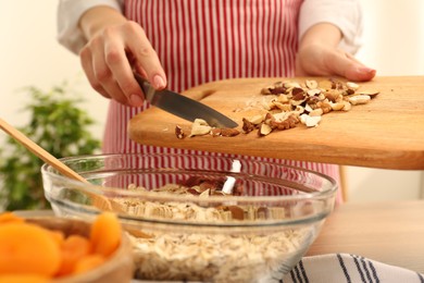 Making granola. Woman adding nuts into bowl with oat flakes at table in kitchen, closeup