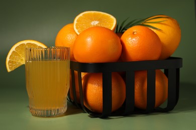 Fresh oranges in metal basket and glass of juice on green background, closeup