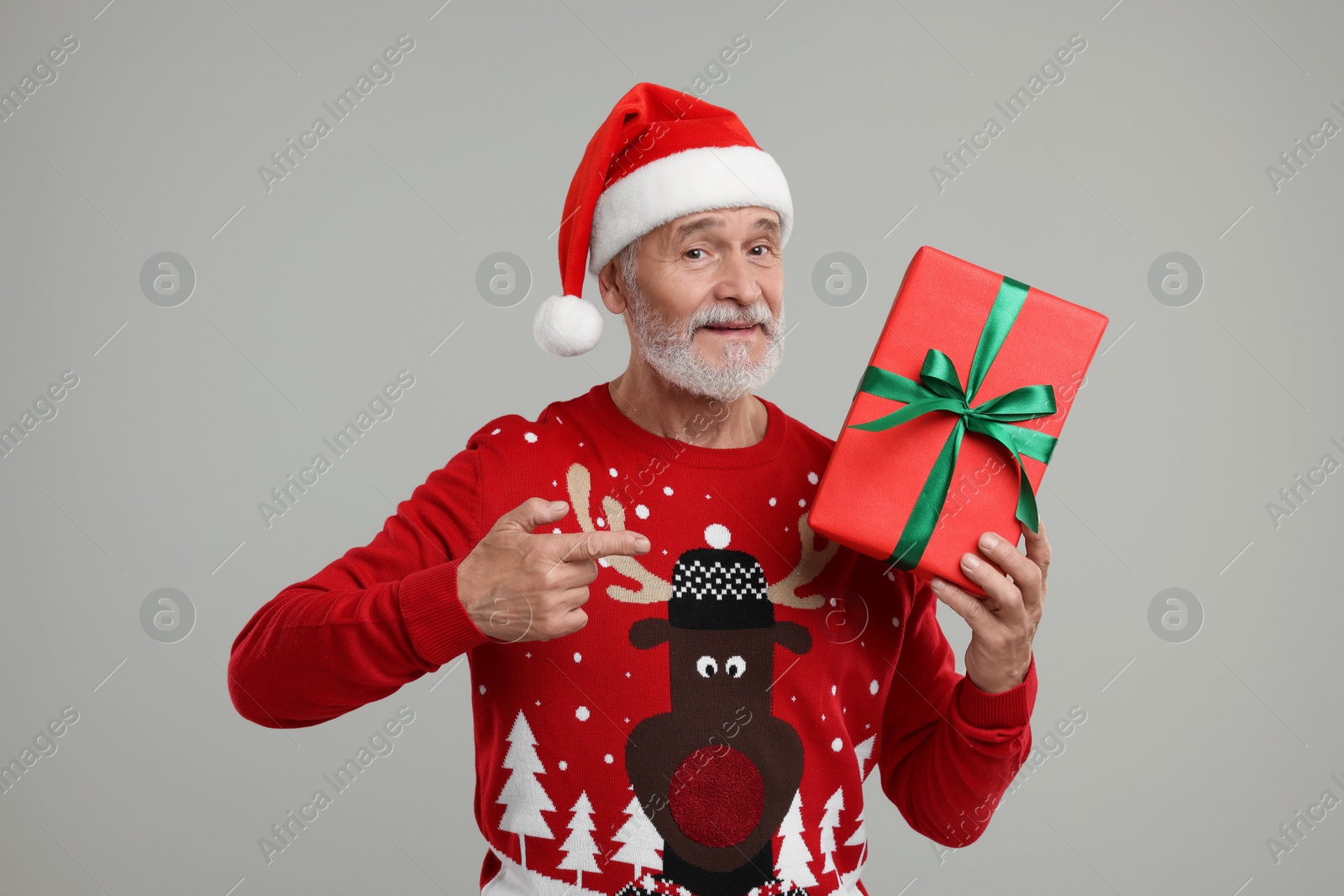 Photo of Senior man in Christmas sweater and Santa hat holding gift on grey background