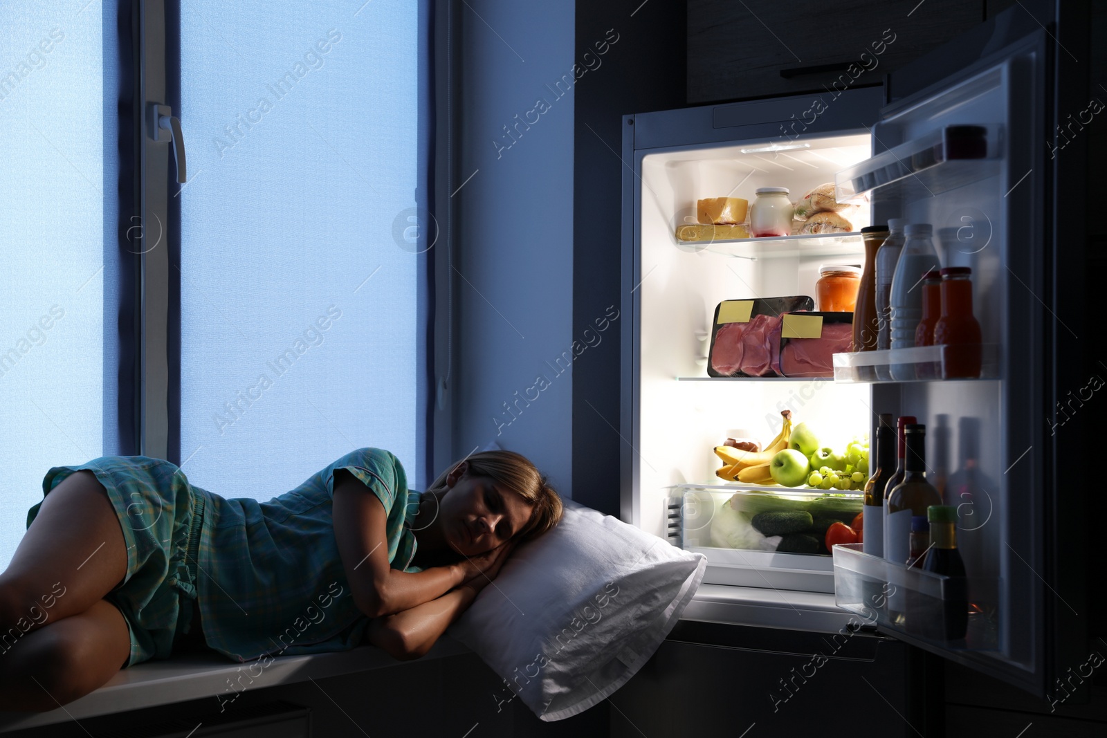 Photo of Woman sleeping on window sill near open refrigerator in kitchen at night