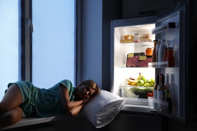 Photo of Woman sleeping on window sill near open refrigerator in kitchen at night