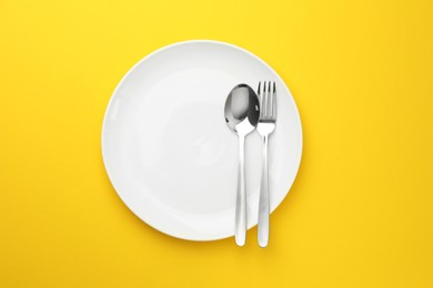 Photo of Clean plate, fork and spoon on yellow background, top view