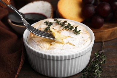 Photo of Eating tasty baked camembert with fork from bowl at wooden table, closeup