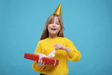 Photo of Happy little girl in party hat opening gift on light blue background