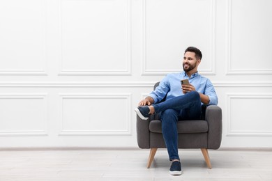 Photo of Handsome man with smartphone sitting in armchair near white wall indoors, space for text