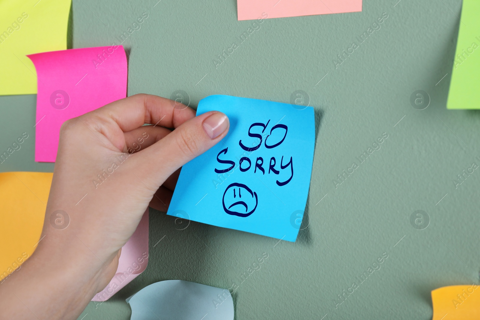 Image of Apology. Woman sticking paper note with text So Sorry and drawn sad face onto grey wall, closeup